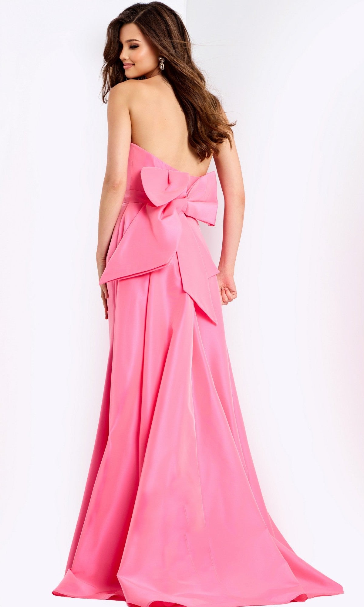 A woman models the Jovani 47495, a strapless, floor-length pink prom dress with a large back bow, standing against a plain white background.